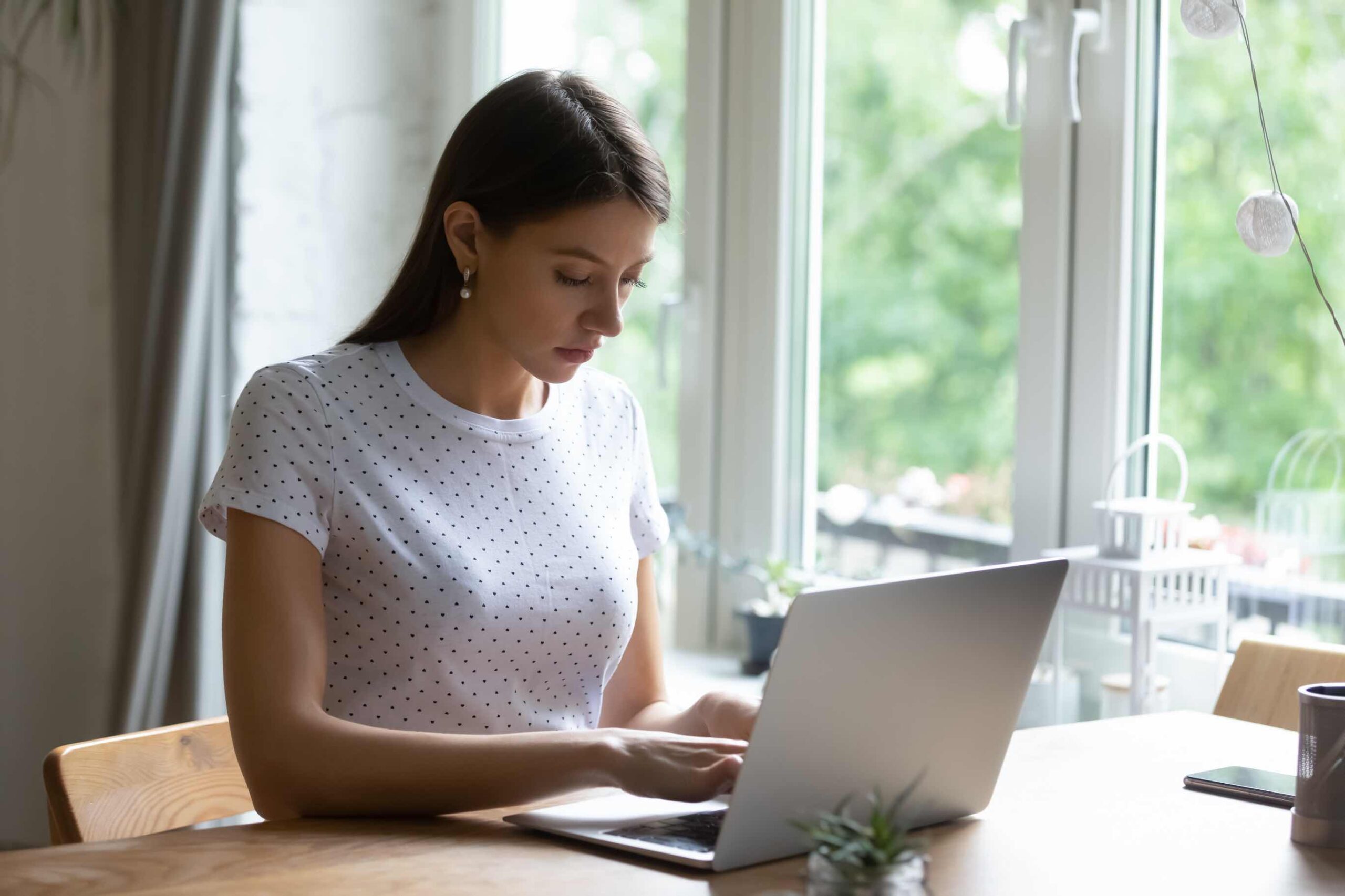 Uma mulher a escrever num computador portátil junto a uma janela.