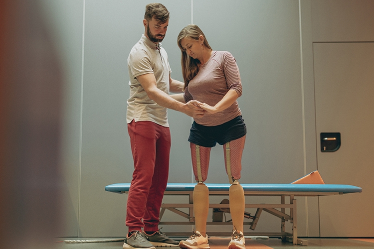 A man helping a woman stand on two prosthetic legs.