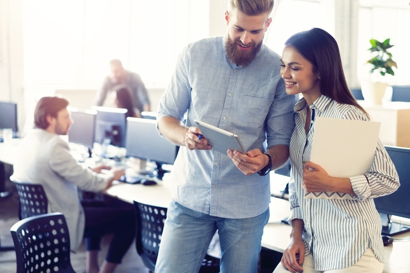 A man showing a woman something on a tablet inside an office with other workers.